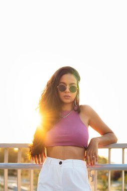 Güneş gözlüklü genç bir Latin kadının gün batımında çekilmiş bir portresi. Kameraya bakıyor ve poz veriyor.