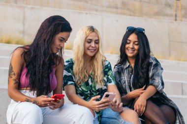 Üç genç, çok ırklı arkadaş açık havada bir merdivende oturmuş akıllı telefon kullanıyor ve eğleniyorlar.