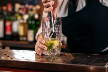Kadın barmen kokteyl barında uzun bardakta mojito hazırlıyor. Kireç ve nane yapraklarını havan topu ile ezer.