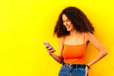 Sarı duvara yaslanmış cep telefonunu kullanan şık bir afro kadın.