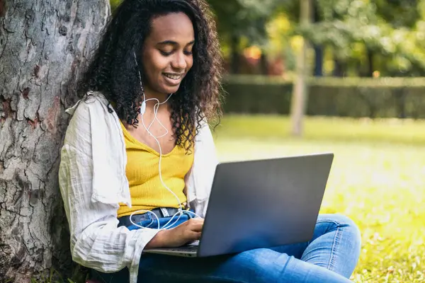 Sarı tişörtlü Afro-Amerikalı kız kampüs çimlerinde oturmuş dizüstü bilgisayarla üniversite ödevi yapıyor. Kulaklıkla müzik dinleyip ders çalışıyor gibi görünüyor.