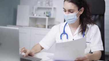 Steril maskeli genç bayan doktor covid 19 salgını sırasında dizüstü bilgisayarla masada oturuyor.