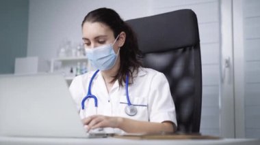 Steril maskeli genç bayan doktor covid 19 salgını sırasında dizüstü bilgisayarla masada oturuyor.