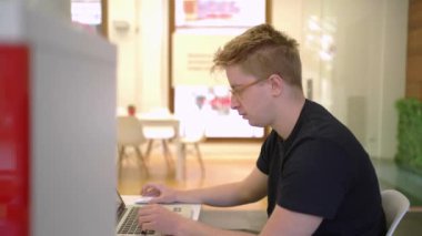 Gözlüklü, masa başında oturmuş, emlak bürosunun internet defteri üzerinde çalışan konsantre bir erkek işçinin yan görüntüsü.