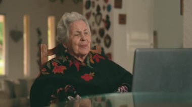 Kısa gri saçlı yaşlı bayanın sağ görüşünü yansıt. Sandalyeye oturup kız kardeşiyle vedalaşırken evde netbook üzerinden video görüşmesi yapıyor. 