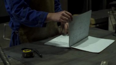 Önlüklü ve iş kıyafetli isimsiz bir erkek işçi. Metal yapıları olan hafif işyerindeki mezuranın yanındaki masada cereyan yapan taslaklar var.