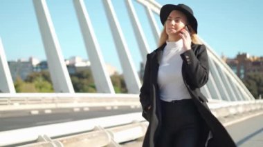 Sırt çantalı ve şapkalı genç sarışın kadın güneşli bir günde asfalt yolda yürürken cep telefonuyla konuşuyor.