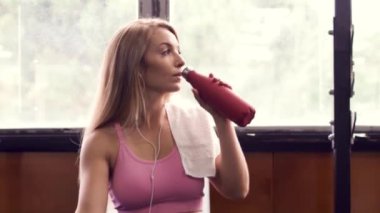 Spor giyim sektöründe atletik kadın kulaklıkla müzik dinlerken ve spor salonunda spor yaparken akıllı telefon kullanırken temiz su içiyor.