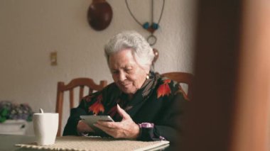 Sıcak giysiler içinde gülümseyen yaşlı kadın tablet ve çay ile masaya oturmuş ekrana bakıyor.