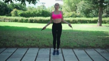 Spor elbiseli, atlama halatıyla atlayan yeşil ağaçlarla parktaki kaldırımda egzersiz yapan bir kadın vücudu.