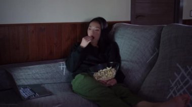 Genç Asyalı kadın gündelik giysiler içinde kanepede oturup evde patlamış mısır yiyor ve dizüstü bilgisayardan film seyrediyor.