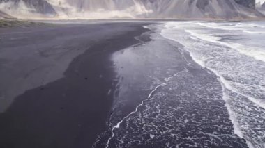 İzlanda 'daki bulutlu gökyüzüne karşı Vestrahorn Dağı' nın yakınlarındaki ıslak kıyılara vuran köpüklü deniz dalgalarının drone görüntüsünü yükselt.
