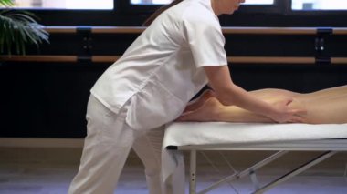 Klinikteki fizyoterapi seansında, beyaz cüppeli, tanınmayan bir masöz, tarladan çıkmış kadın bir hastaya masaj yapıyor.