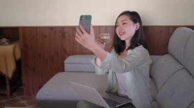 Evde dizüstü bilgisayarla kanepede otururken cep telefonuyla kendi portresini çeken genç etnik bir kadın.