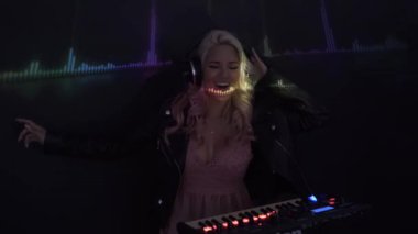 Kulaklıklı mutlu genç bayan DJ gülümsüyor ve gece kulübünde parti sırasında duman içinde dururken müzik çalmak için sentezleyici kullanıyor.