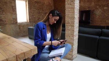 Cep telefonuyla internette sörf yapan uzaktan kumandalı bir kadın bacak bacak üstüne atmış ve dizüstü bilgisayarla çatı katında oturuyor.