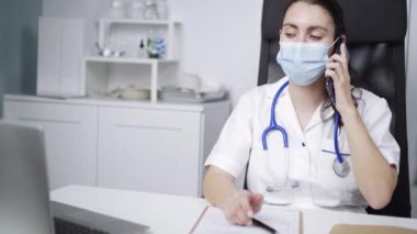 Üniformalı ve koruyucu maskeli kadın doktor Coronavirus salgını sırasında hastanede dizüstü bilgisayarla masa başı notlar alırken cep telefonuyla konuşuyor.