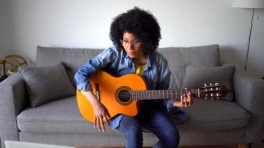 Gülümseyen, kısa kıvırcık saçlı, gözlüklü, rahat koltukta oturup şarkı söyleyen ve dizüstü bilgisayarın önünde gitar çalan Afrikalı Amerikalı bayan müzisyen.