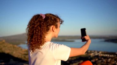 Kıvırcık saçlı kadın yürüyüşçünün, güneşli bir günde denizin üzerindeki kayalık kayalıklarda battaniyede selfie çekerken görüntüsü.