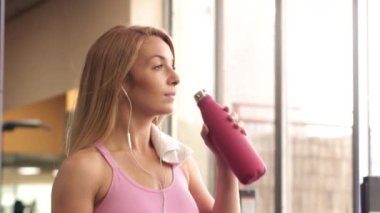 Spor giyim sektöründe atletik kadın kulaklıkla müzik dinlerken ve spor salonunda spor yaparken akıllı telefon kullanırken temiz su içiyor.
