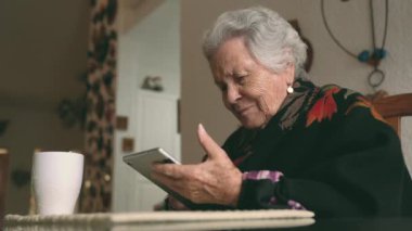 Sıcak giysiler içinde gülümseyen yaşlı bir kadın tablet ve çay bardağıyla masaya oturmuş ekrana bakıyor.