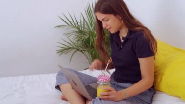 Yatağında bacak bacak bacak üstüne atmış, dizüstü bilgisayar başında oturmuş evde uzak bir projede çalışırken ve meyve suyu içerken pozitif genç bir kadın serbest yazar.