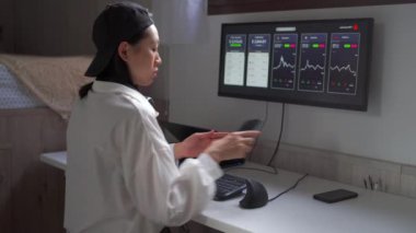 Etnik kadın dağıtıcının bilgisayar monitöründe kripto para birimi grafikleri ve evdeki masada cep telefonu üzerinde çalışmasının arka planı