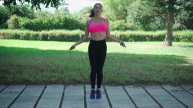 Spor elbiseli, atlama halatıyla atlayan yeşil ağaçlarla parktaki kaldırımda egzersiz yapan bir kadın vücudu.