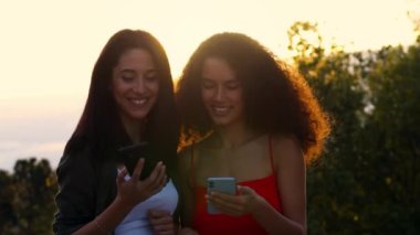 İki genç kadın oturuyor ve cep telefonundan bir şeyler izliyorlar.