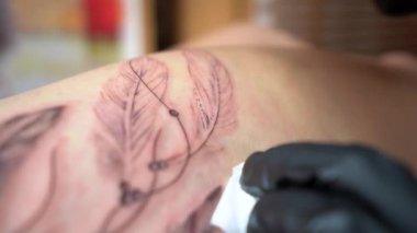 Profesyonel makineli eldivenli tanınmayan bir dövmeci. Stüdyodaki müşterinin vücuduna yaprak şeklinde dövme uyguluyor.