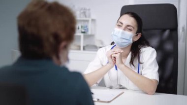 Koruyucu yüz maskesi takmış, elinde dosya kağıdıyla kadın doktor salgın sırasında birbirine bakarken hastanede kimliği belirsiz yaşlı bir kadınla konuşuyor.