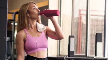 Spor giyim sektöründe atletik kadın kulaklıkla müzik dinlerken ve spor salonunda spor yaparken akıllı telefon kullanırken temiz su içiyor.