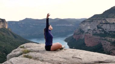 Uzun siyah saçlı, spor giyimli, uzun boylu, kararlı genç bayan sporcu kayalık vadide antrenmandan önce ısınırken yan eğilip esneme hareketleri yapıyor.