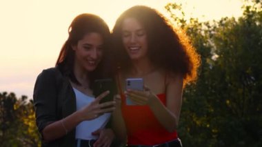 İki genç kadın oturuyor ve cep telefonundan bir şeyler izliyorlar.