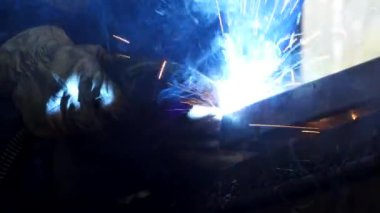 Eldiven ve üniformalı yüzsüz işçi fabrika inşaatlarının yanındaki masada metal kaynakçılığı yapıyor.