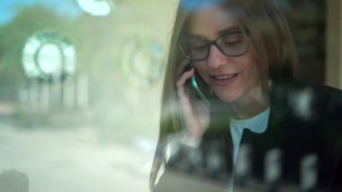 Camdan, gözlüklü, uzun düz saçlı ve resmi giyinmiş bir kadın kahve dükkanında otururken akıllı telefondan konuşuyor.