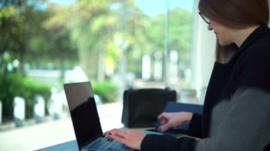 Gözlüklü, gözlüklü, akıllı telefon kullanırken laptopta çalışan kadın girişimcinin yan görüntüsü