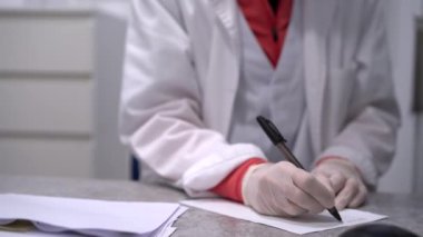 Lateks eldivenli, üniformalı, tanınmayan bir kadın doktor. Masada oturuyor ve reçeteyi kağıda yazıyor.