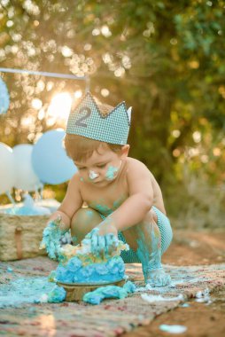 Küçük tatlı bir çocuk dışarıda ikinci yaş gününü berbat bir pasta parçasıyla kutluyor. Altında bez ve kağıt bir taç var. Etrafı balonlarla çevrili mavi pastayı neşeyle eşeliyor.