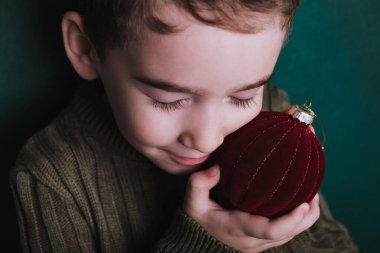 Gözleri kapalı genç bir çocuk, huzurlu tatil sıcaklığını ifade eden kırmızı kadife bir Noel süsünü nazikçe kucaklıyor..