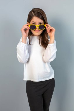 Neşeli genç bir kız, aşırı büyük sarı güneş gözlükleriyle gri bir arka plana karşı neşeli bir şekilde poz verir.