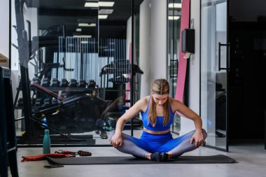 Mavili bir atlet, bir spor salonundaki esneklik rutinine konsantre olmak için mindere oturmuş bir esneme hareketi yapıyor.