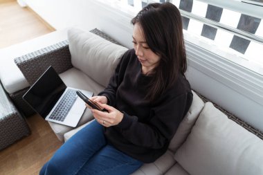 Bir kadın dizüstü bilgisayarı açık bir şekilde kanepeye oturur, dikkatle akıllı telefonuna bakar, rahat bir uzaktan çalışma ortamı yaratır.