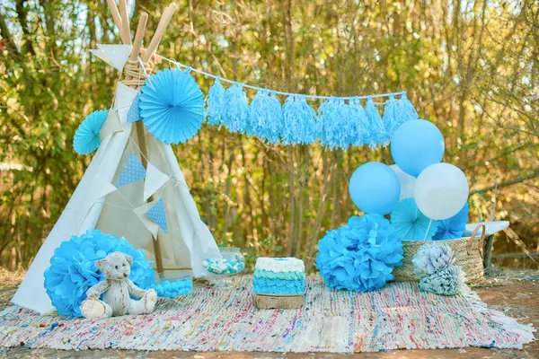 stock image A whimsical outdoor setup for a celebration, featuring a white teepee adorned with sky-blue paper fans, matching balloons and pompoms, and a cozy seating area with a textured rug