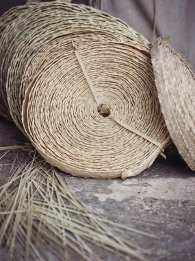 Özenle dokunmuş palmito sepetleri, titiz bir spiral tasarımı içerir, kırsal beton zeminde çiğ palmito liflerinin yanında sergilenir. Görüntü, eski sepet dokuma sanatını vurgular.
