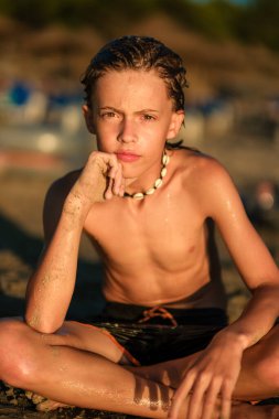 Düşünceli, ıslak, deniz kabuğundan kolye takan, yaz tatili boyunca kumlu sahilde oturan ve kameraya bakan çocuk.