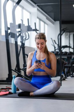 Mavi spor kıyafetli bir kadın akıllı telefonunu dikkatle kullanır. Muhtemelen spor programını takip eder ya da antrenman programını planlar.