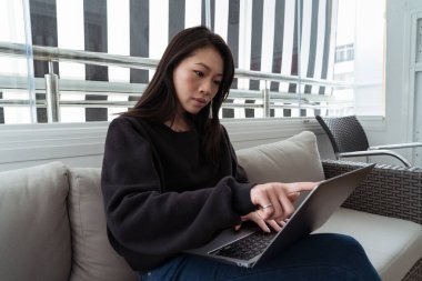 Odaklanmış genç bir kadın, rahat bir şekilde koltukta otururken dizüstü bilgisayar kullanır, modern çizgili perde arkası...