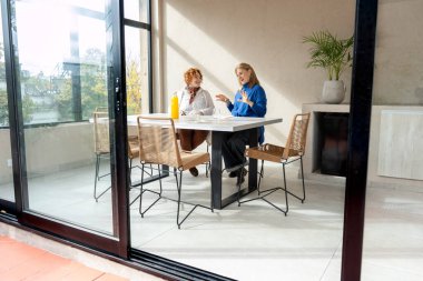 İki orta yaşlı kadın modern güneşli bir balkon ortamında şık beyaz bir masada otururken çağdaş mobilyalar ve sakin bir manzarayla çevrili.