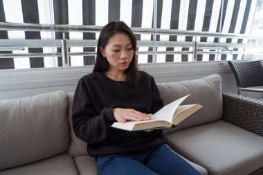 Genç bir kadın pencere kenarındaki bir kanepede panjurlarla rahatça otururken kitap okumaya dalmış ve evin içinde huzurlu bir ortam yaratıyor.